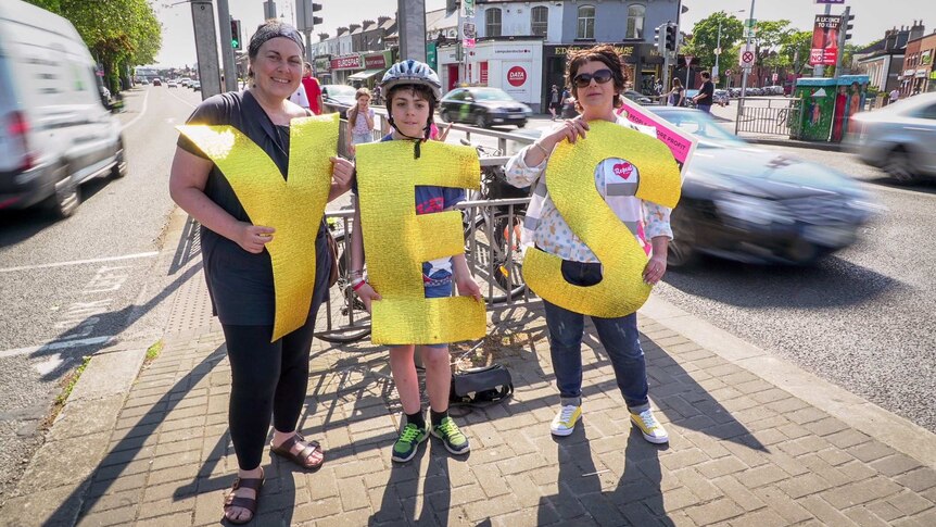 Two women and a young boy hold big gold letters spelling "YES" at an intersection.