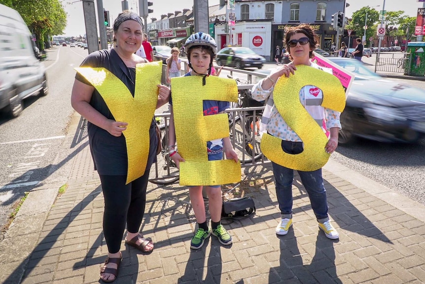 Two women and a young boy hold big gold letters spelling "YES" at an intersection.