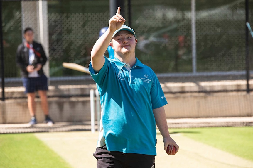 A young man with a disability holds up a finger in the 'howzat' gesture. He stands in cricket nets, holding a six-stitcher.