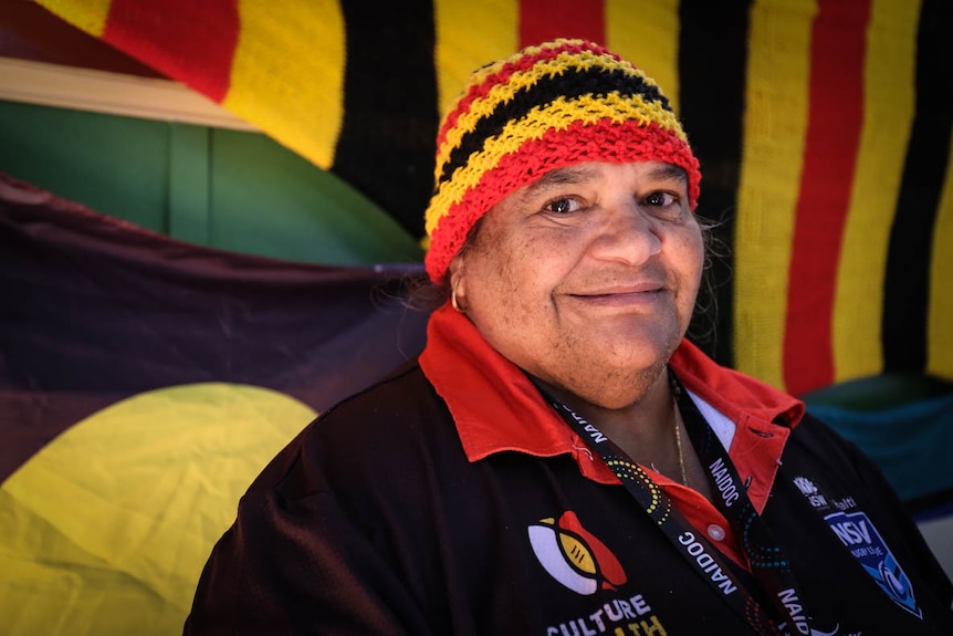 An Indigenous woman wearing a rad, yellow and black crocheted hat, in front of an Aboriginal flag.