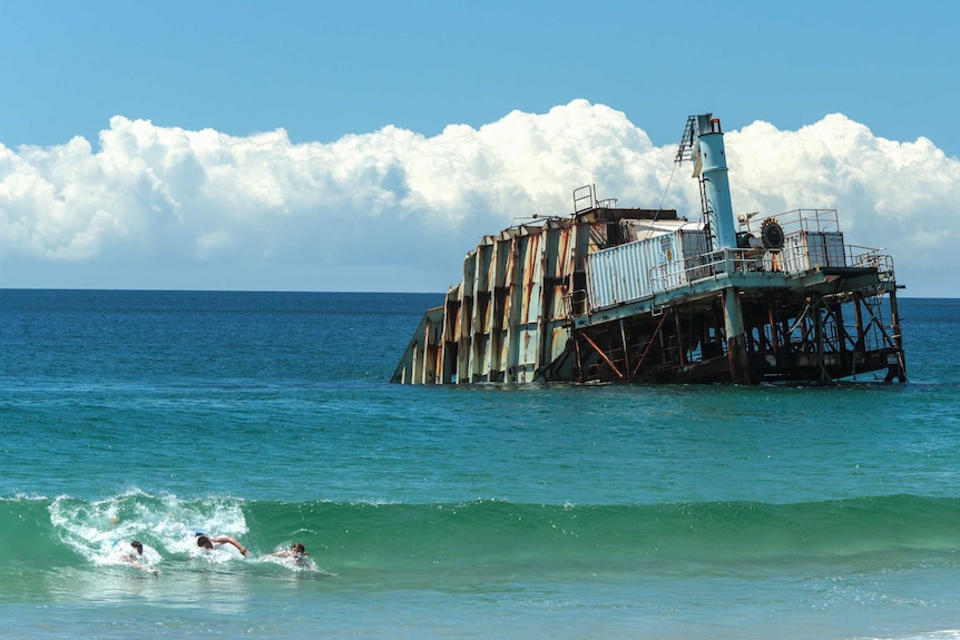 A huge, rusted machine in the ocean with kids playing in the surf.