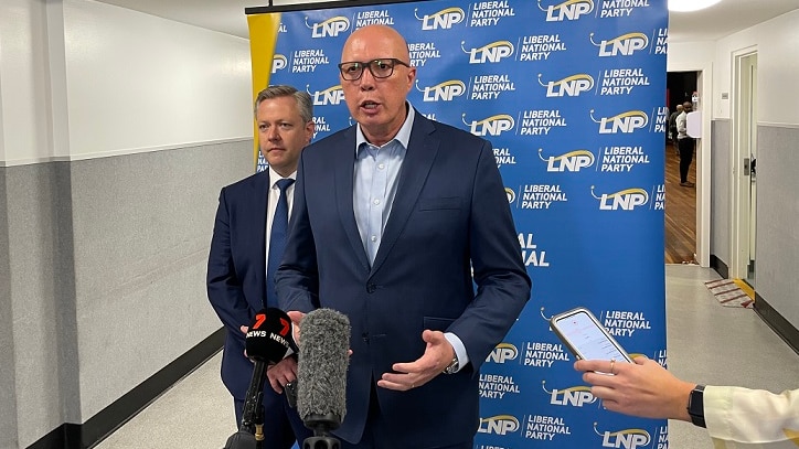 A man speaking to the press with another man behind him in front of a Liberal Party banner.