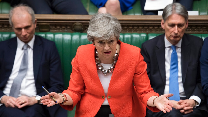 Theresa May wearing a bright reddish orange jacket, with her hands outstretched and a concerned look on her face.