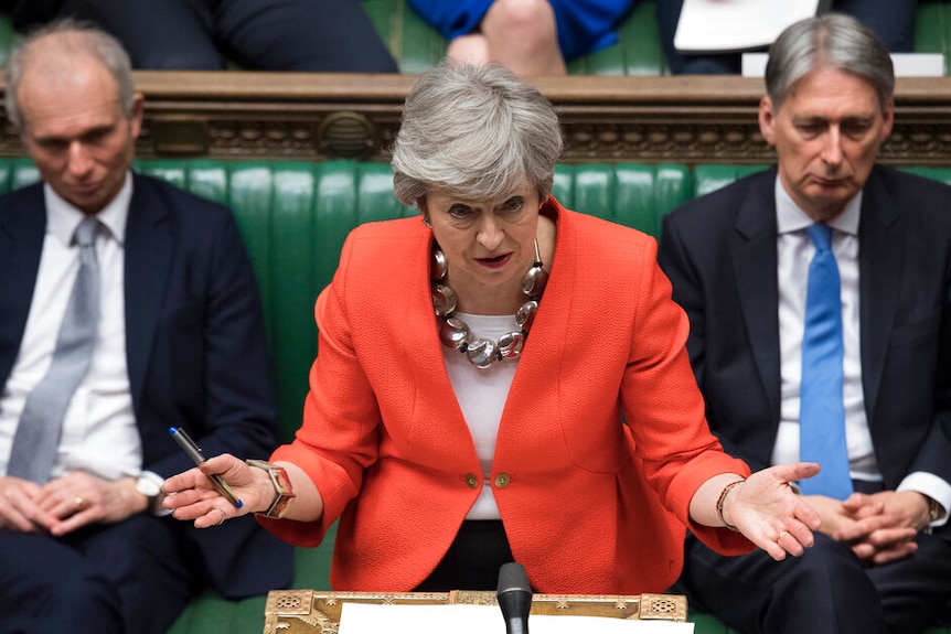 Theresa May wearing a bright reddish orange jacket, with her hands outstretched and a concerned look on her face.