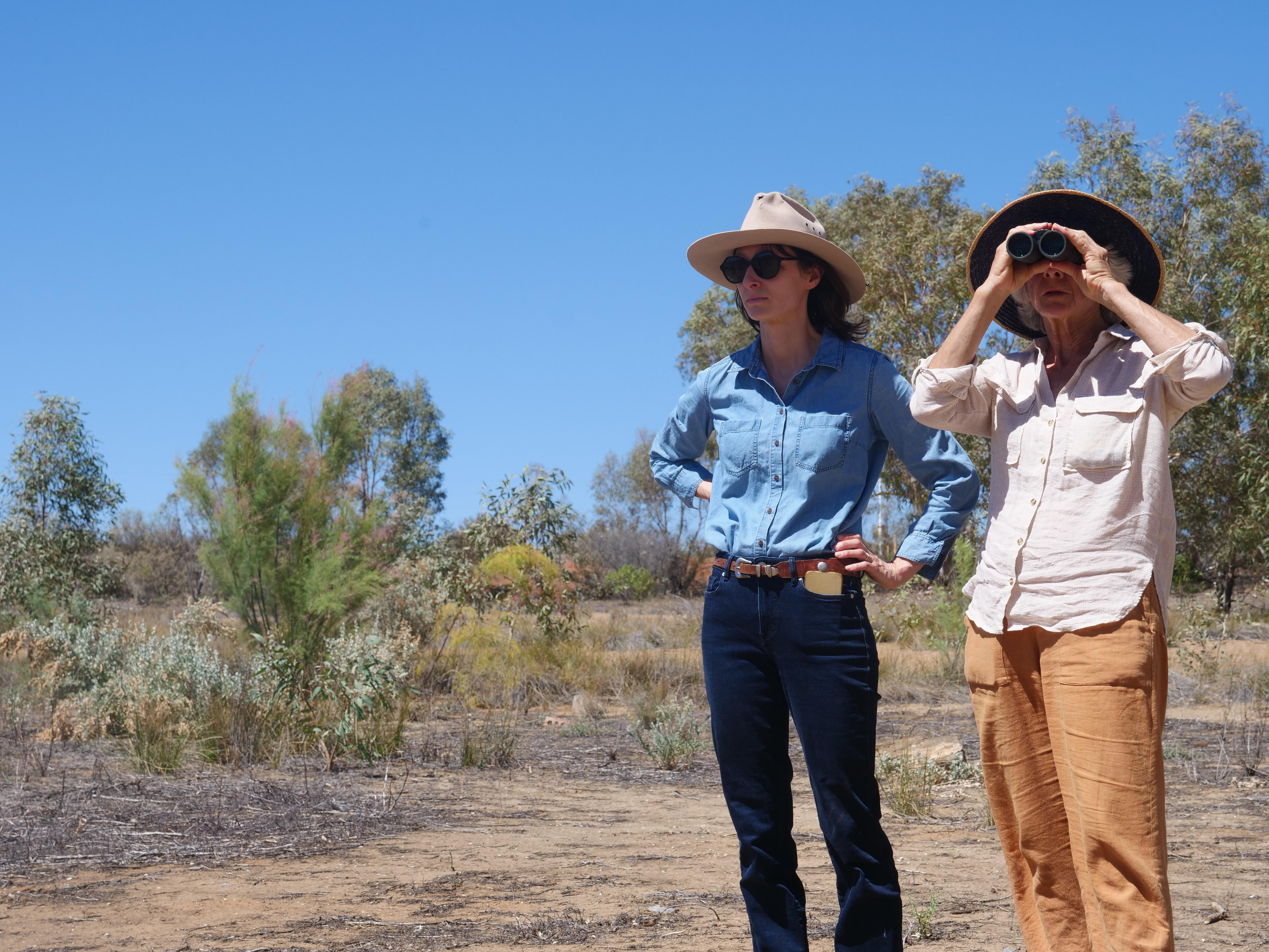 Anika está com as mãos nos quadris nas margens do seco Lago Oriental, ao lado de sua mãe, que observa através de binóculos.