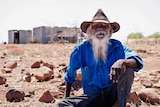 A photo of Indigenous man Paddy Doolak sitting on the ground with the old Wave Hill station in the background.