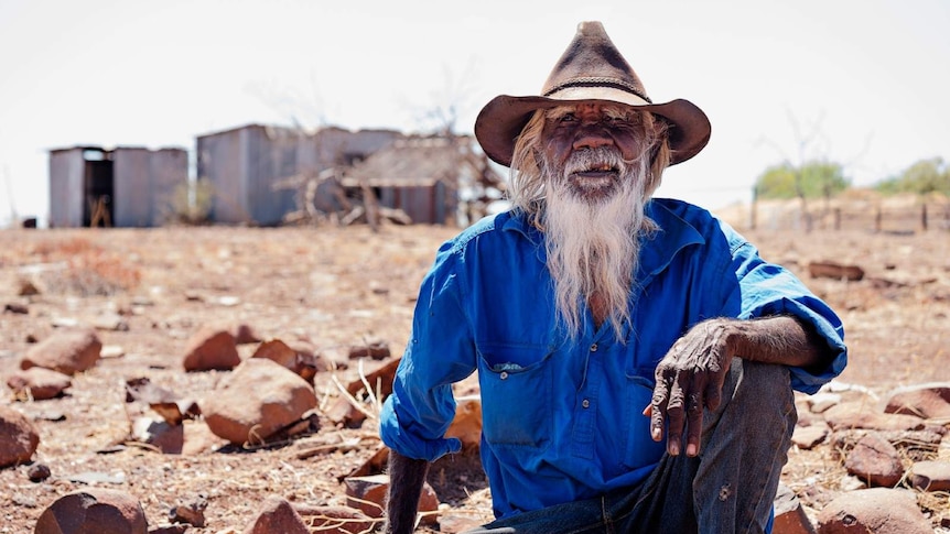 A photo of Indigenous man Paddy Doolak sitting on the ground with the old Wave Hill station in the background.