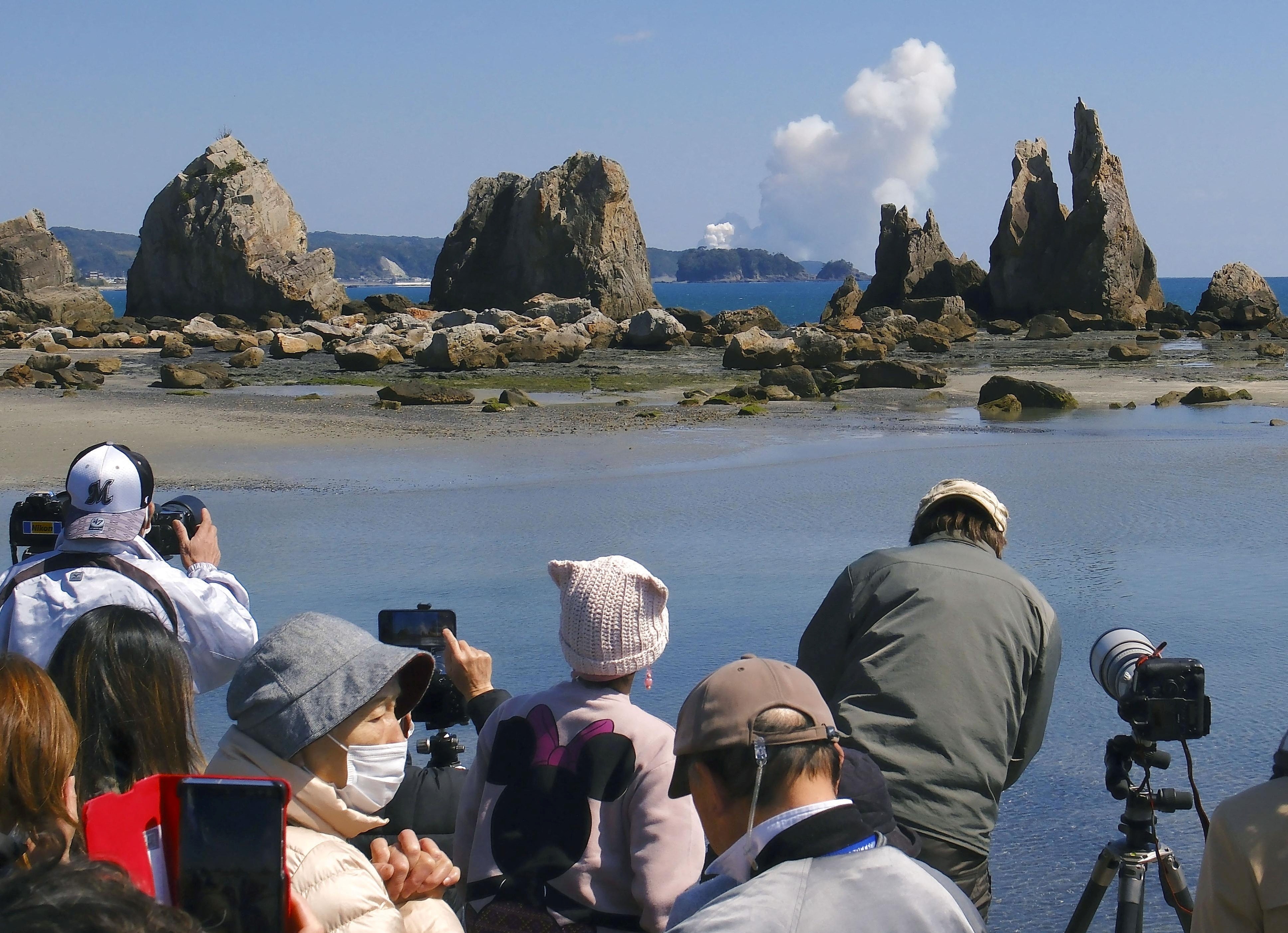 A group of Japanese people watch and take photos of a smoke cloud rising in the distance across a body of water.