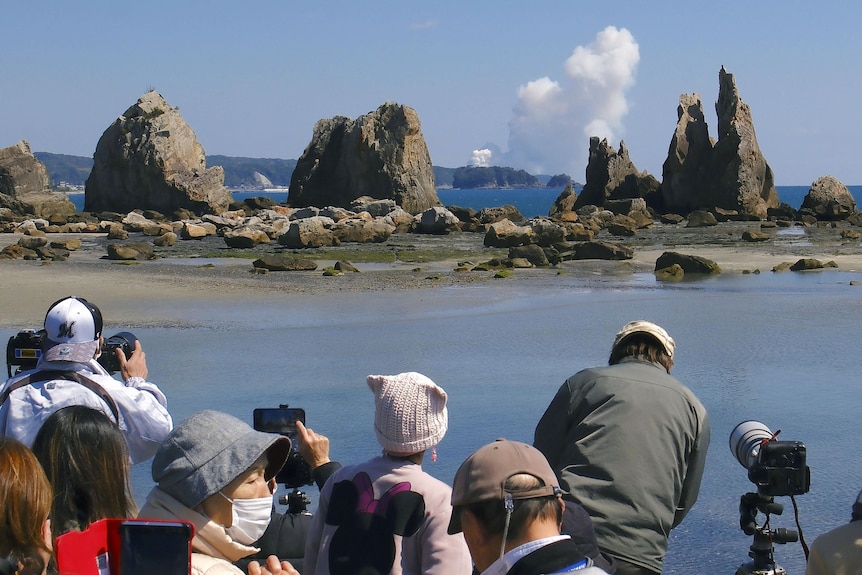 A group of Japanese people watch and take photos of a smoke cloud rising in the distance across a body of water.