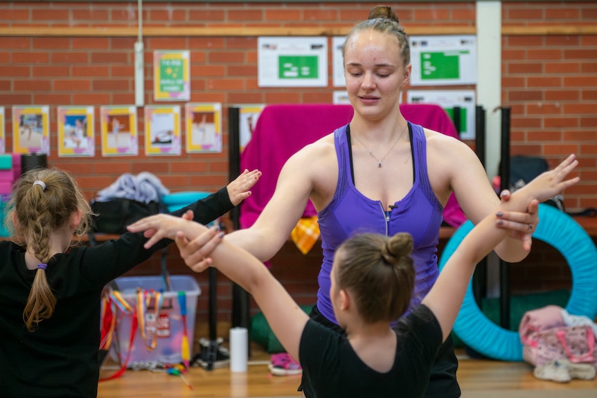 A young athletic woman with hair pulled into a tight bun gently corrects the hand placement of her younger gymnastics student