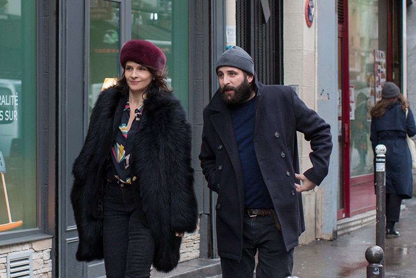 A smiling woman with fur hat and man with beanie, both in dark jeans and coats, walk on shopfront street sidewalk in the day.
