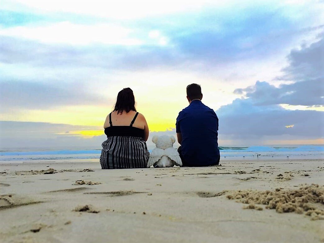 A man and woman sitting on the beach with their backs to the camera.  A bear with angel wings is between them.