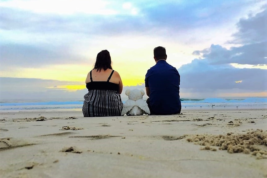 A man and woman sitting on the beach with their backs to the camera.  A bear with angel wings is between them.