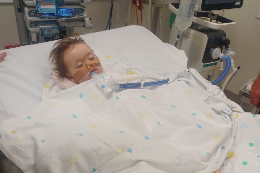 Little girl lies in hospital bed with feeding and breathing tube in her mouth. 