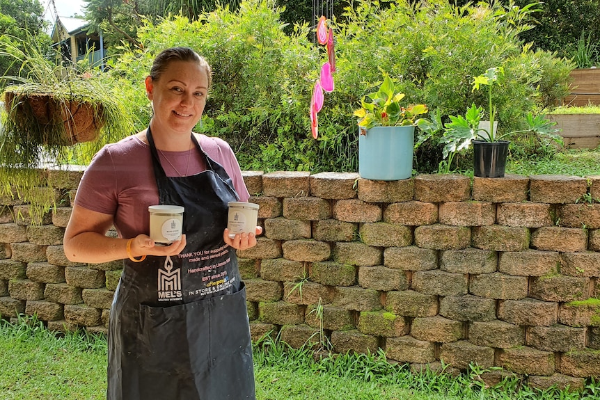 A woman stands in a garden wearing a black work apron and holding two candles.