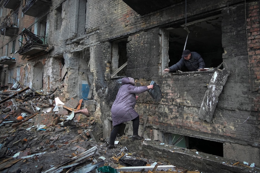 People gather their belongings from a damaged house, with one passing items out a destroyed window to another.