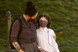 A woman wearing a mask puts her arm around a young girl who is holding a dog leash outside and also wearing a mask.