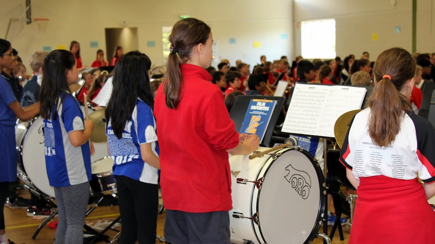Students face the band conductor at a band rehearsal inside a school hall.