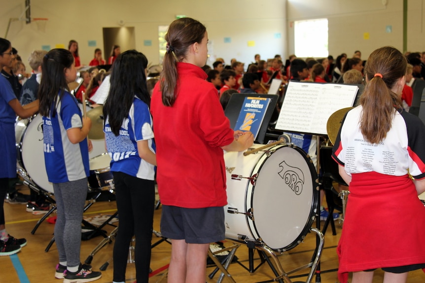 Students face the band conductor at a band rehearsal inside a school hall.