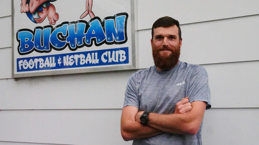 A man with a scrubby beard standing with his arms crossed in front of the sign for the Buchan Football Netball Club.