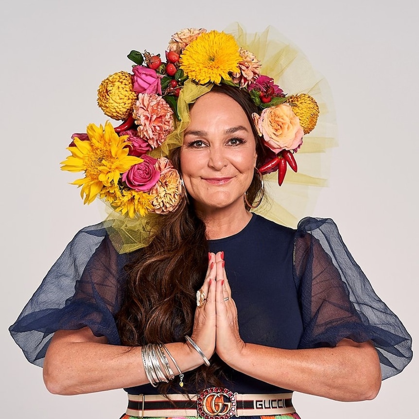 A woman stands with her hands in a praying pose, wearing a headpiece covered in flowers.