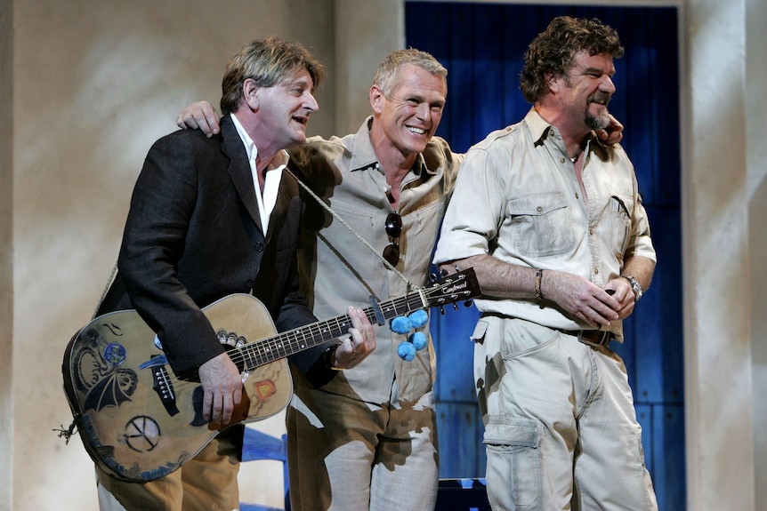 Three men on a stage, one holding a guitar, with arms around eachother 
