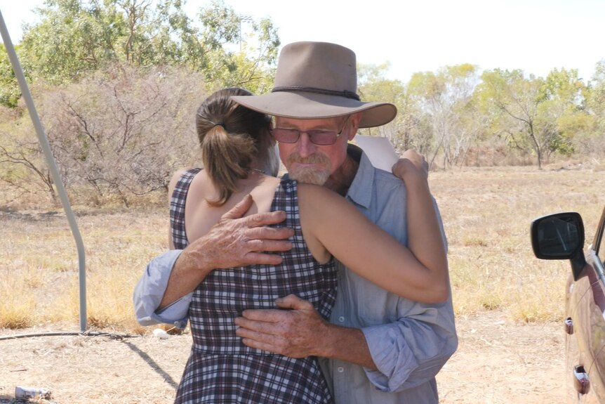 two people hugging, she is in a dark, checked, sleeveless dress and he is in a pale blue shirt and Akubra hat