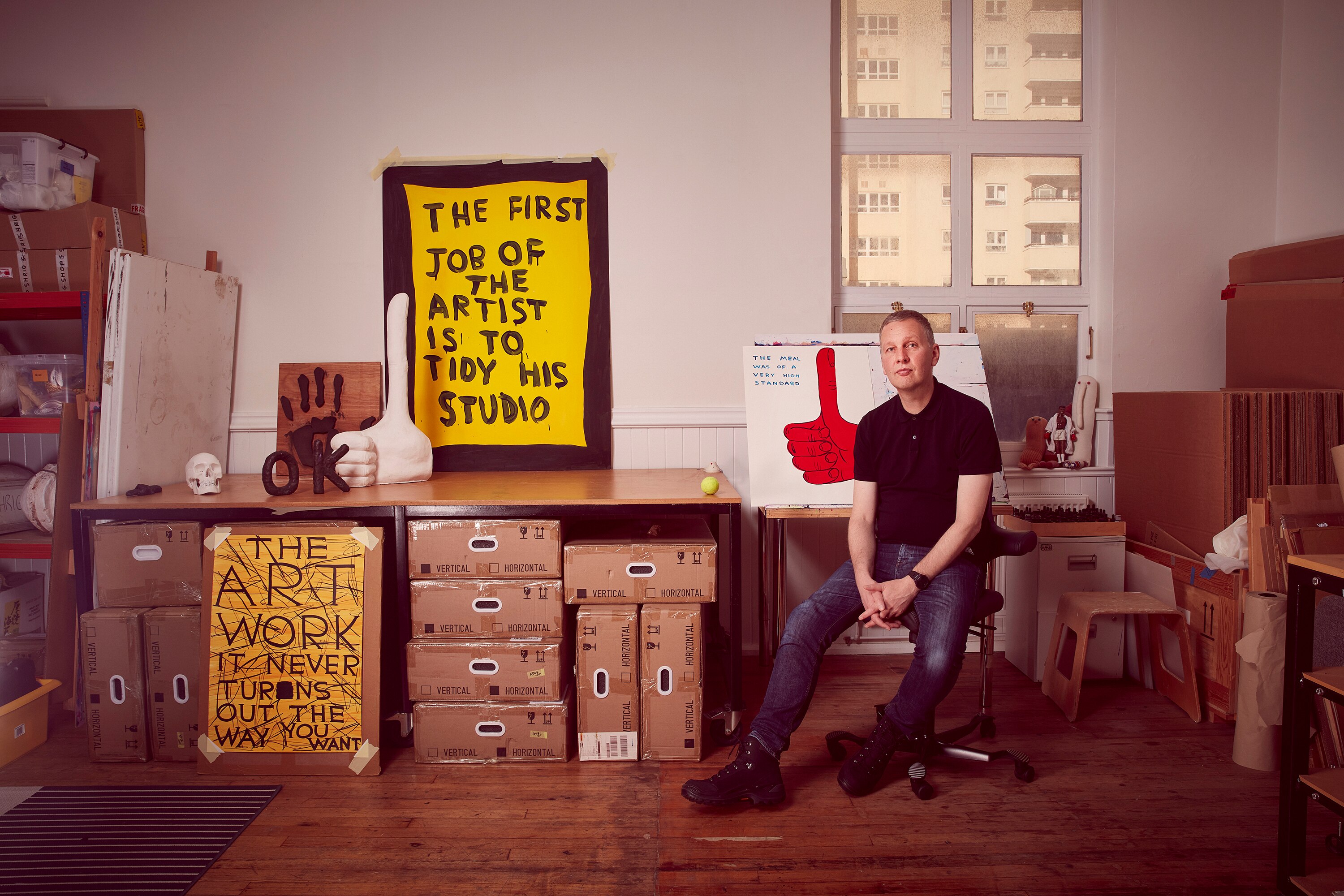 Artist David Shrigley sits in his studio in front of boxes and paintings.
