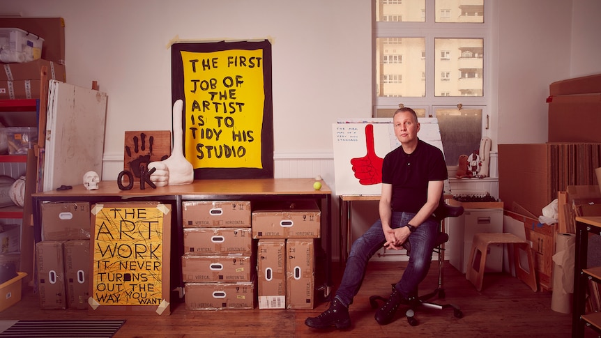 Artist David Shrigley sits in his studio in front of boxes and paintings.