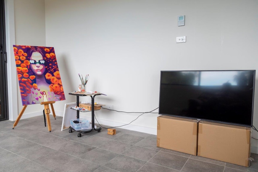An easel with a bright purple and orange painting sits in a sparse living room. A TV is on two cardboard boxes.