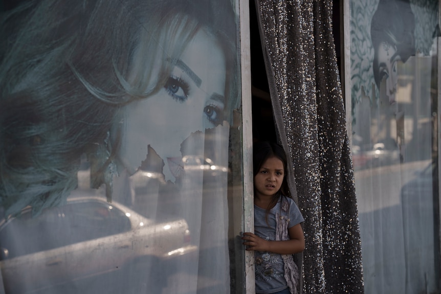 A young girl stand near a shop window with a torn poster of a woman. 