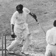 Bradman looking back at his stumps with the bails off with the wicket keeper and slip behind him.