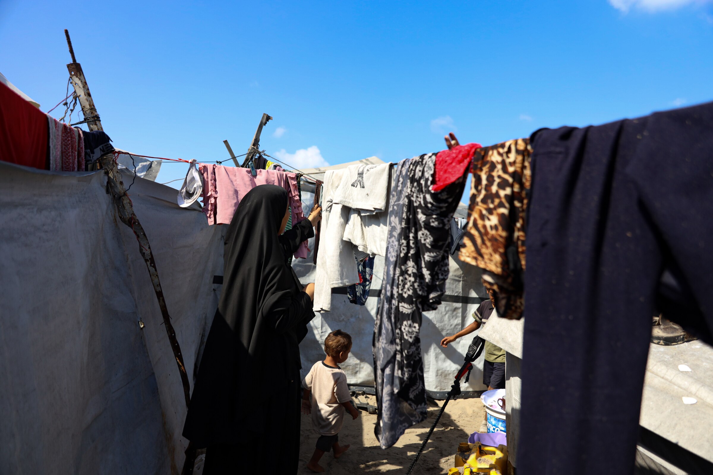 A woman wearing a hijab hanging cloths on a line with a child walking below