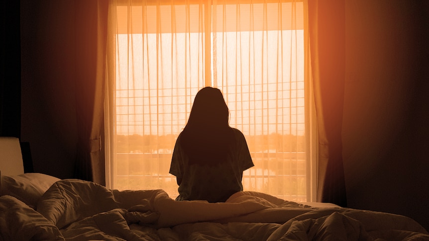 Silhouette of woman sitting on bed facing window.
