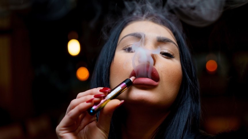 A woman holds a vape to her lips and smoke pours from her mouth