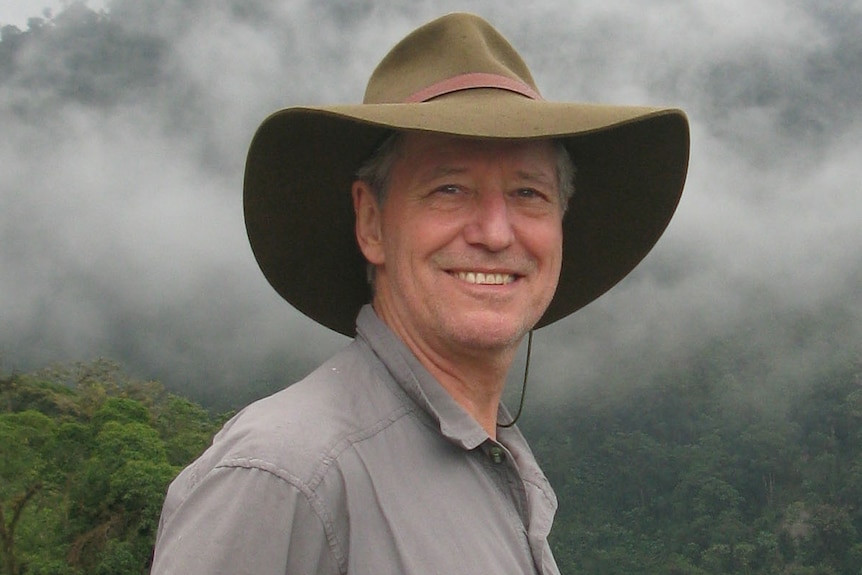 A man in a full-brimmed hat stands close to the camera with rainforest in the distant back ground