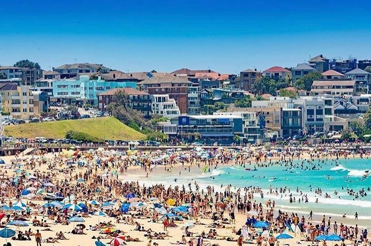 Thousands of people on the sand at a beach and swimming in the water, with housing development in the background.