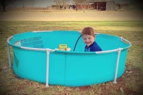 A young boy with red hair sits in a small pool smiling at the camera.