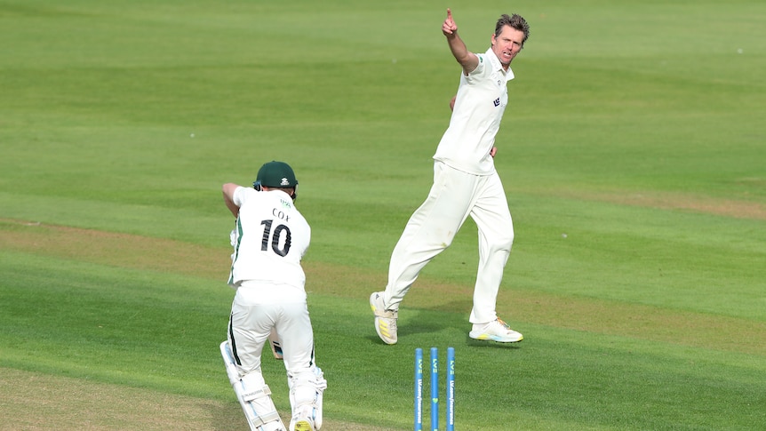 A bowler turns away while pointing to the sky as he runs to celebrate as a batsman stands with the bails off behind him.