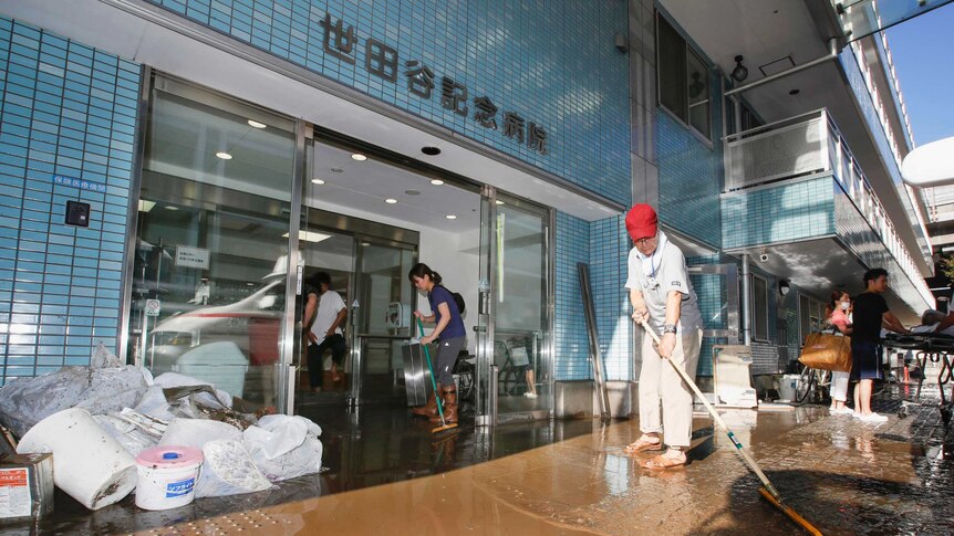 People use brooms and mops to clean up muddy water at the entrance of a blue tiled building.