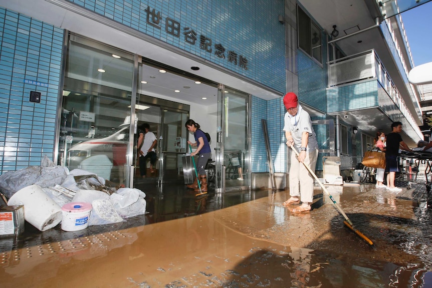 People use brooms and mops to clean up muddy water at the entrance of a blue tiled building.