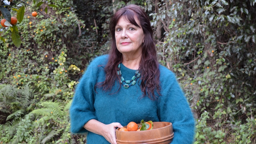 A portrait of a woman  with long brown hair with fringe stands in a garden holding a bowl of oranges