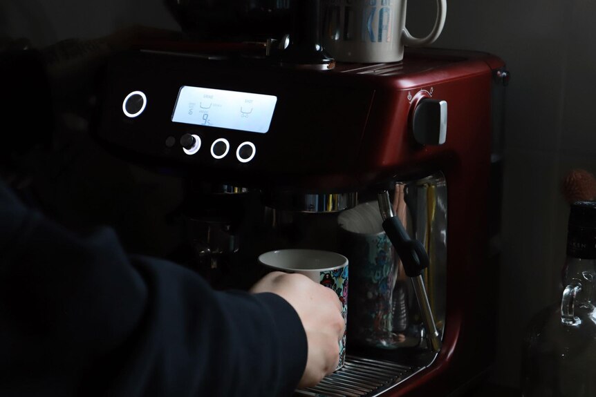 Жена с тъмен пуловер държи чаша кафе под червена кафе машина.