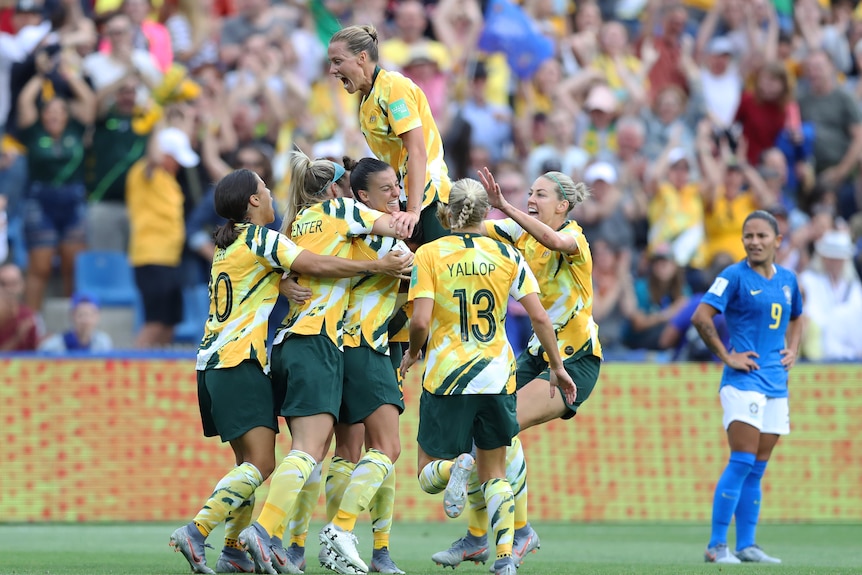 The Matildas huddle around Chloe Logarzo and celebrate while a Brazil player looks on.
