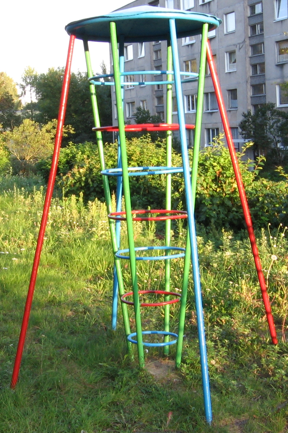 A colourful rocket shaped climbing gym stands in an overgrown field in front of a grey apartment block