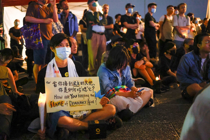 People sit at a protest, with one woman holding a sign saying, "How are YOU keeping the flame of democracy alive?"