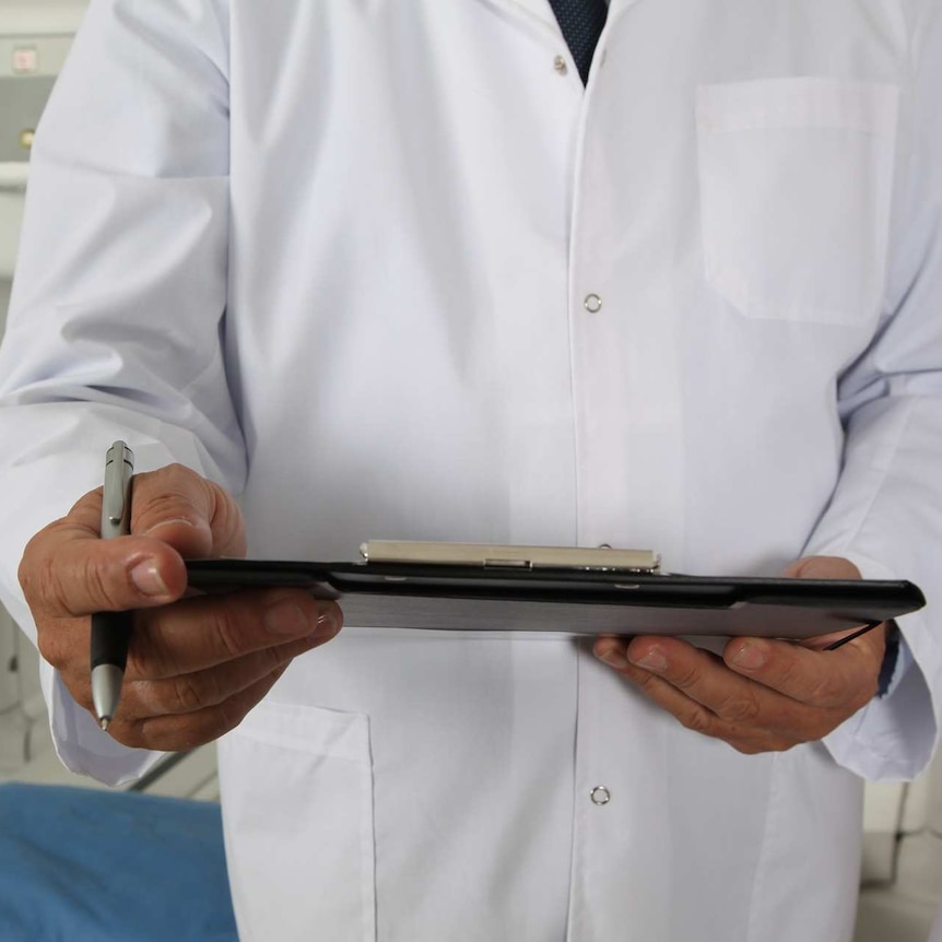 A person in a white doctor coat with a clipboard with a tomograph machine in the background.