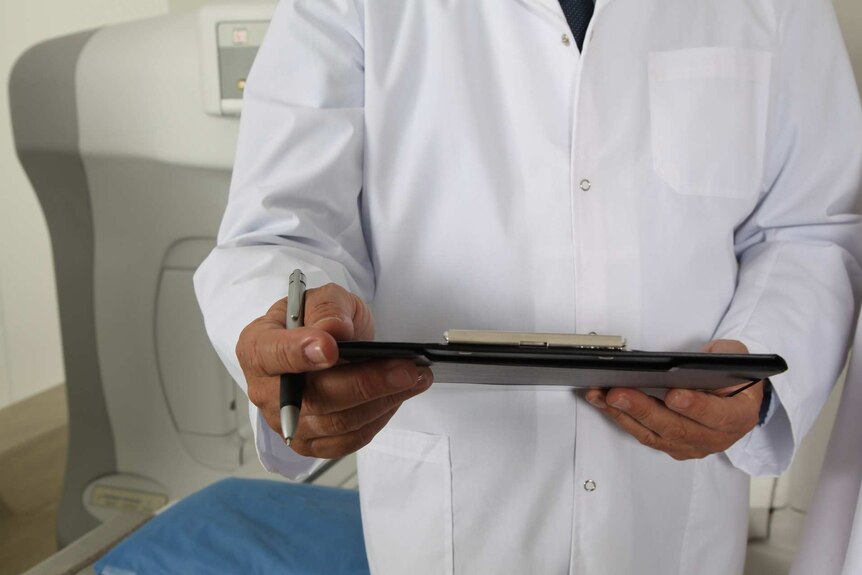 A person in a white doctor coat with a clipboard with a tomograph machine in the background.