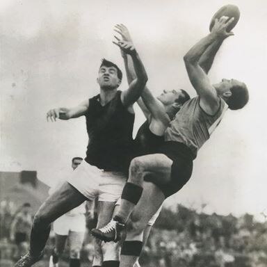 A SANFL player at the back of a pack reaches his arms out abov e his head to grab the ball while two defenders collide.