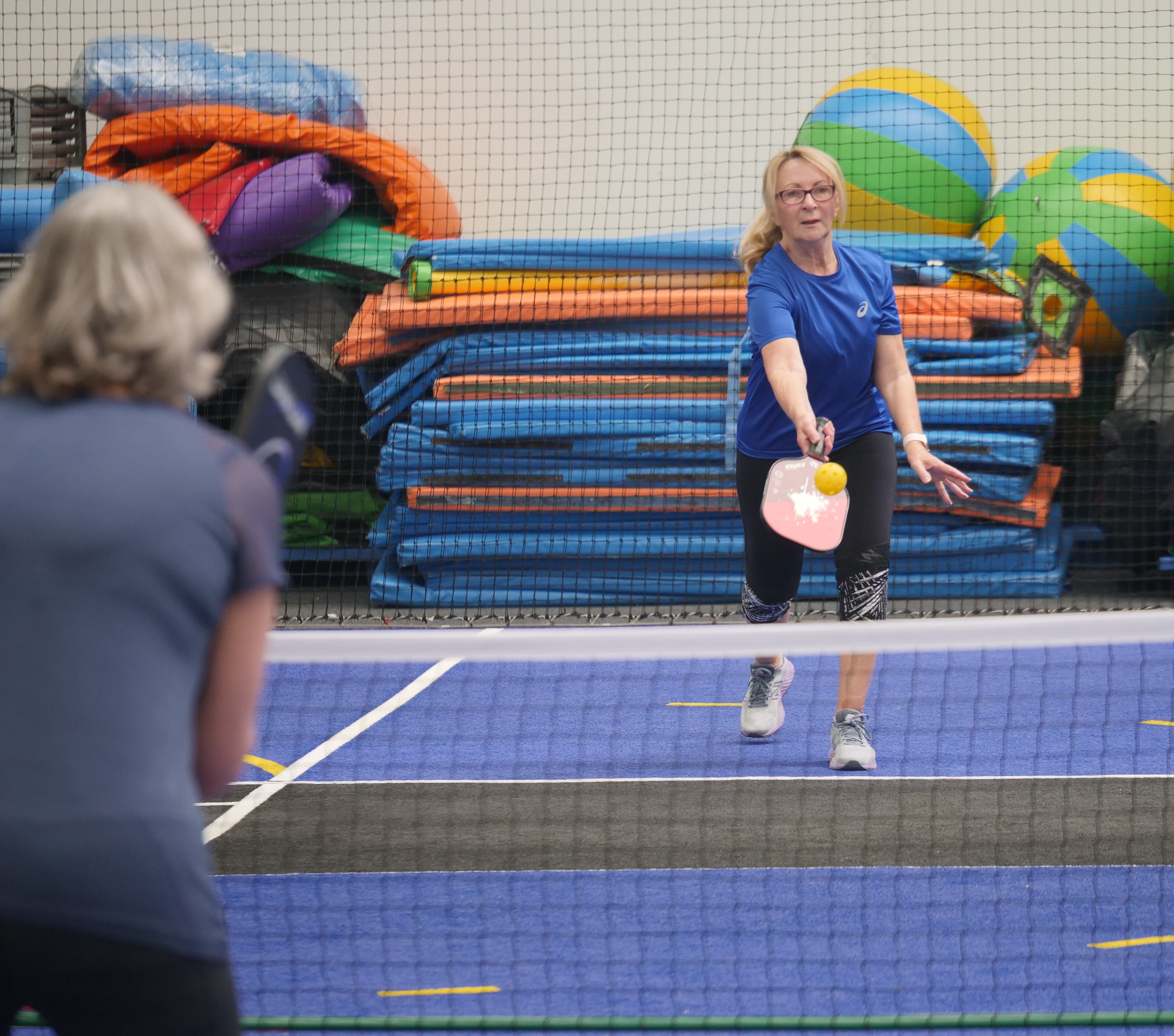 A woman in a blue shirt, hitting a yellow ball over a net with a paddle. She is playing pickleball against an opponent. 